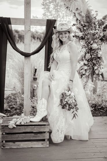 Bride in a lace wedding dress and cowboy hat smiles outdoors with a bouquet and boots.