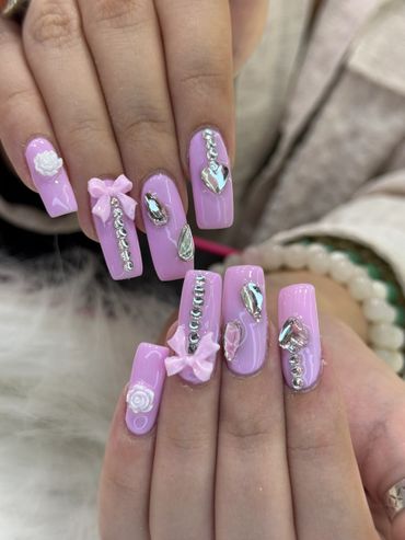 Lavender nails with silver gems, pink bows, and white floral accents.