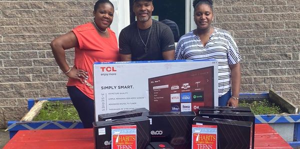 Three people posing with a TCL TV and multiple boxed items on a table outdoors.