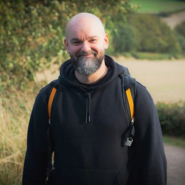 Smiling bald man with a beard outdoors wearing a black hoodie and backpack.
