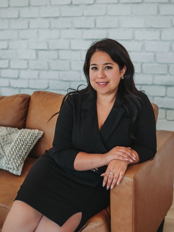 Confident woman in a black dress sitting on a brown couch with a smile.