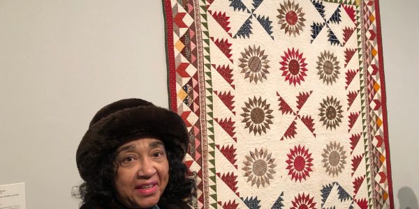 Woman in a brown hat standing near a colorful quilt.