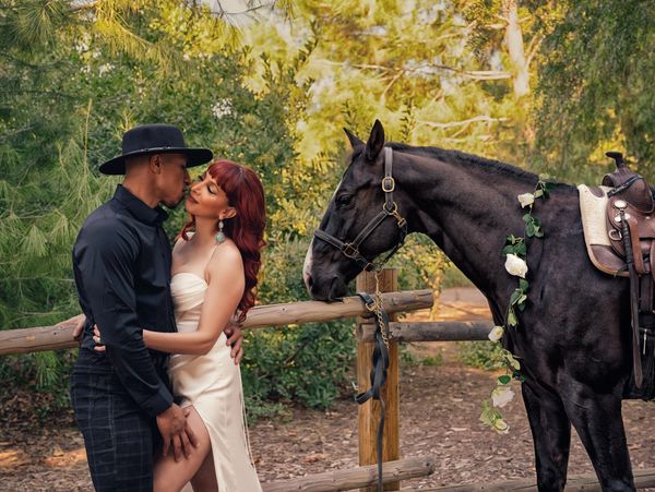 Couple sharing a tender moment beside a decorated black horse in nature.