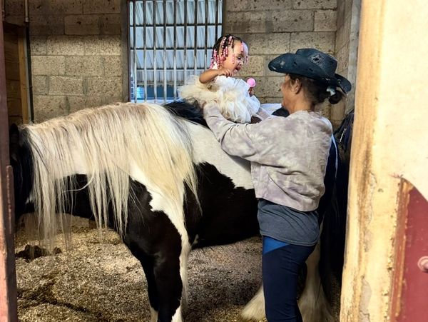 A woman helps a small child sit on a black and white horse inside a stable.