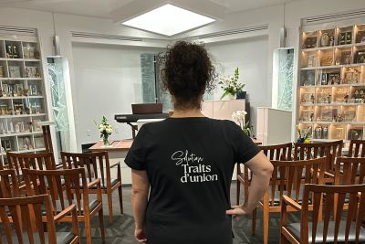 Person wearing a black shirt stands in a small chapel with wooden chairs and shelves.