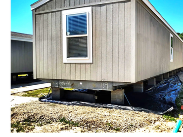 A beige mobile home elevated on concrete blocks with a car parked nearby.