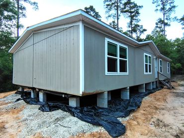 A raised grey house on concrete blocks surrounded by black plastic sheeting and dirt.