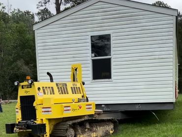 A yellow tracked vehicle moving a white mobile home across grass.