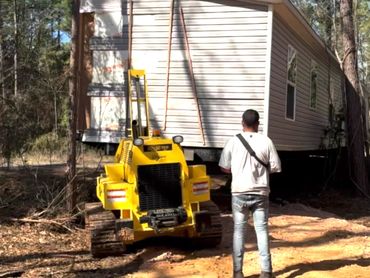 Man stands near yellow machinery moving a small house in a wooded area.