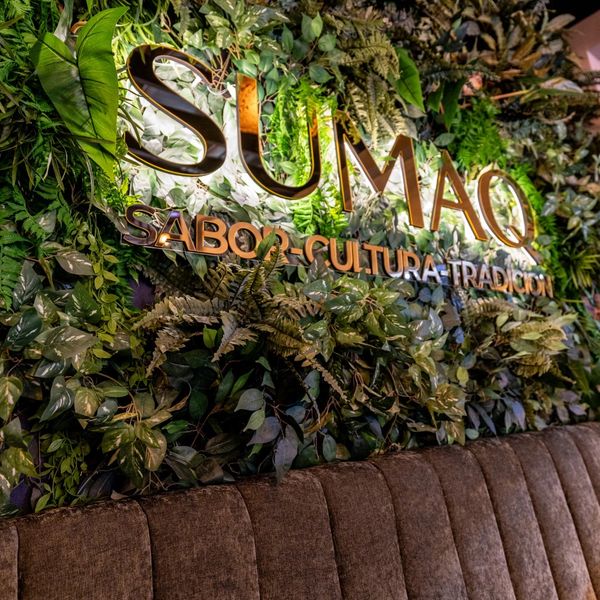 Illuminated SUMAQ sign with greenery and cushioned seating below.
