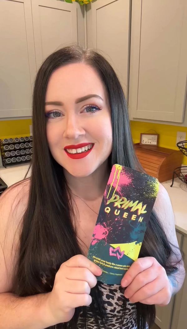 Woman with long dark hair smiles, holding a colorful Primal Queen product package in a bright kitchen.