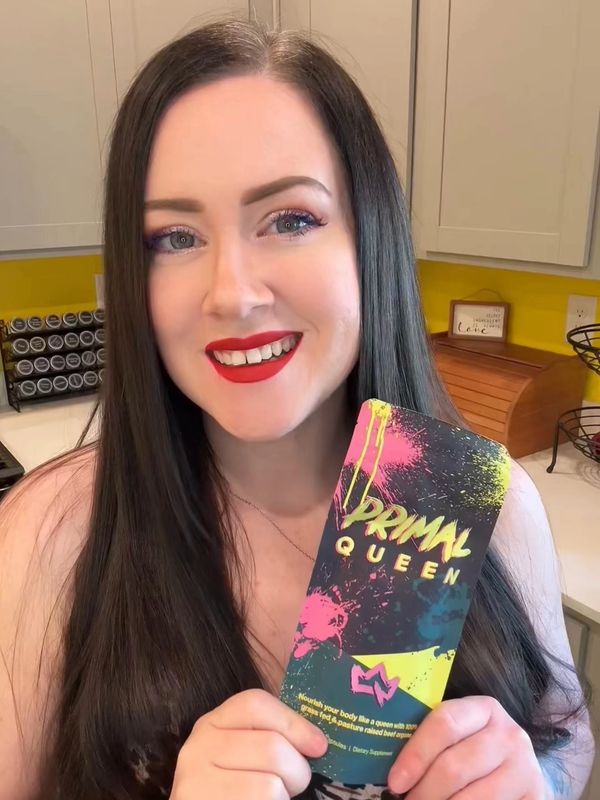 Woman with long dark hair smiles, holding a colorful Primal Queen product package in a bright kitchen.
