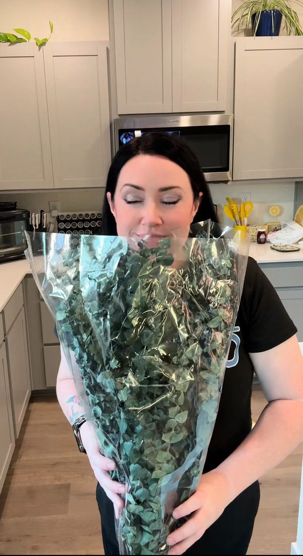 Woman holding a large bouquet of green foliage, smiling with eyes closed in a kitchen.