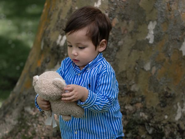 A toddler in blue striped shirt holding a teddy bear near a tree.