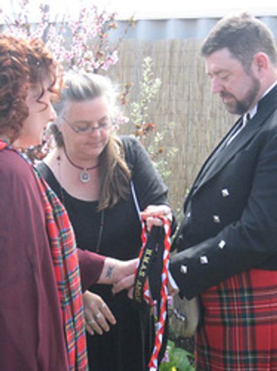 A High Priestess wraps handfasting cords around a couple's clasped hands at their wedding.