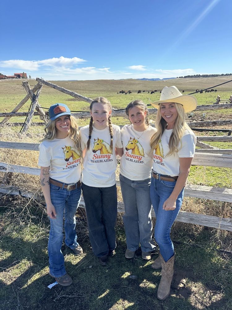 Four smiling women in matching Sunny Trailblazers shirts pose outdoors by a rustic fence.
