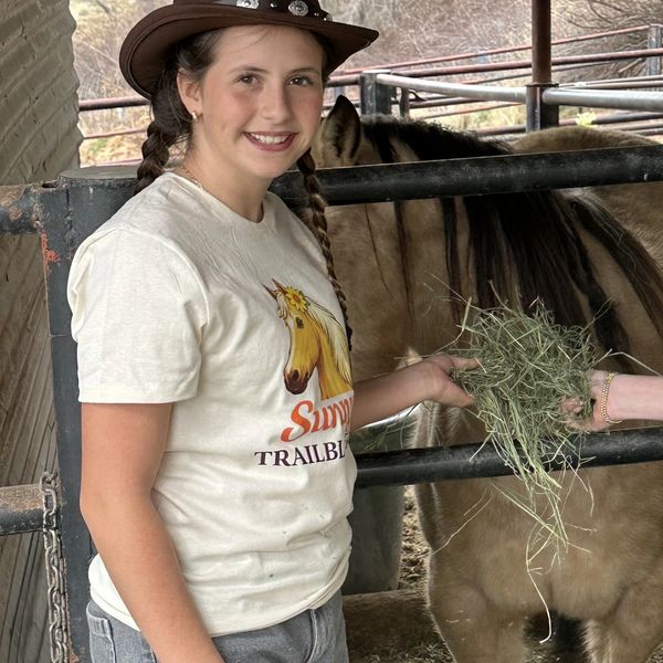 A smiling girl in a cowboy hat feeds a horse hay in a stable.