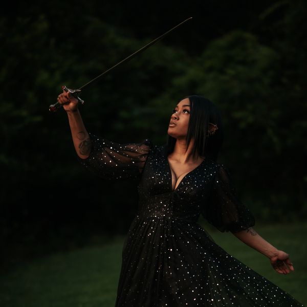 Woman in a sparkling black dress holding a sword aloft in a dark outdoor setting.