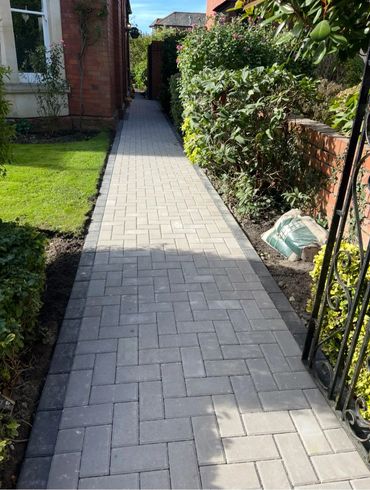 A neat paved pathway bordered by plants and a brick wall leading to a house.