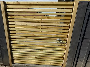 A wooden slat gate with a metal ring handle between black painted walls.