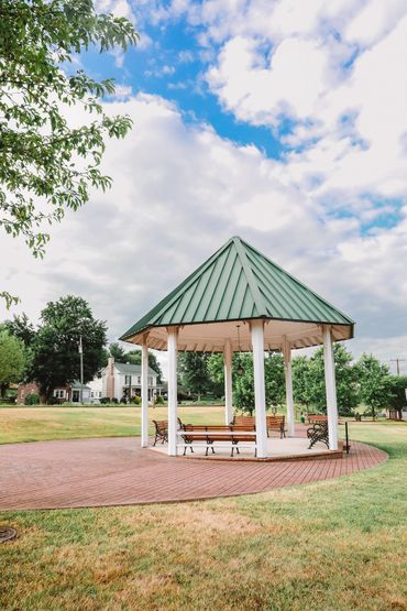 A green-roofed gazebo with benches in a park under a partly cloudy sky.