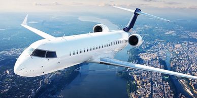 A white aircraft flying over a scenic cityscape and river, with blue skies and clouds.
