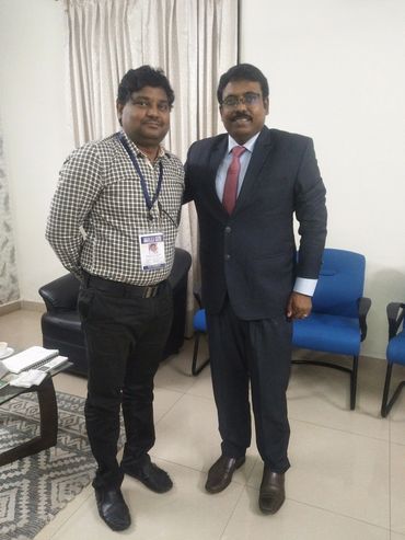 Two men posing together indoors, one in a suit and the other in a checkered shirt with an ID badge.
