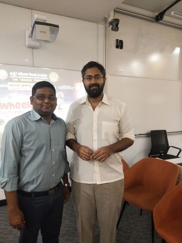 Two men standing in a classroom or seminar room with chairs and a projector screen behind them.