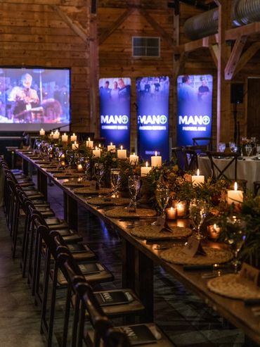 Elegant candlelit dinner setup in a rustic wooden hall with MANO banners and a projection screen.