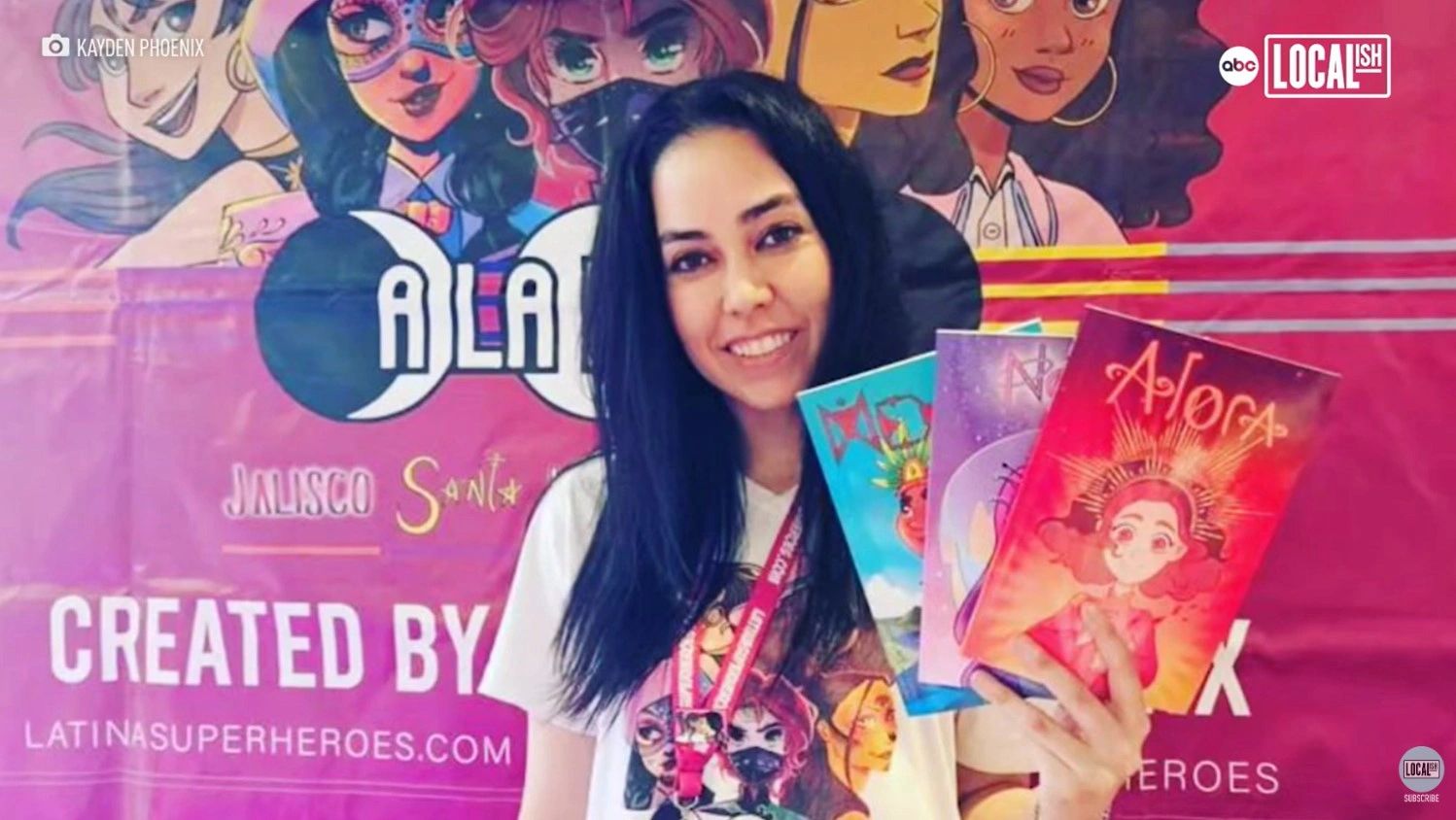 A woman holds colorful comic books in front of a Latina superheroes banner.
