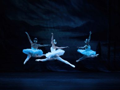 Three ballerinas leap gracefully on stage in blue lighting.