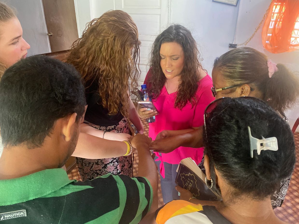 A group of people holding hands in a circle, one woman reading from a phone.