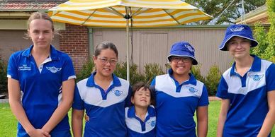 Five kids in blue sports uniforms standing barefoot on grass with a yellow-striped umbrella.