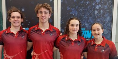 Four young swimmers in matching red shirts posing together indoors.