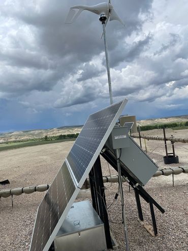 Solar panels and a wind turbine under a cloudy sky in a rural area.