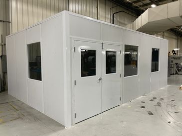 A white modular office with several windows & a double door, installed inside an industrial setting