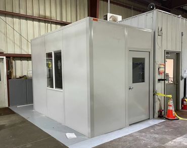 A grey modular prefab with a sliding glass window, door. Inside a warehouse setting.