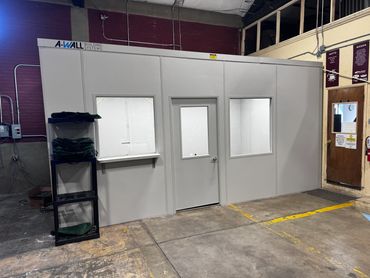 A prefab modular building with a couple windows, door, lights on. installed inside a warehouse cor