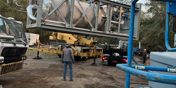 Workers oversee lifting a large industrial tank using a crane.