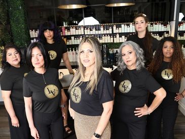 Seven women wearing matching black shirts with a gold logo, posing confidently indoors.