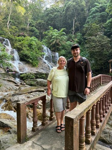 Two people posing on a bridge near a lush waterfall in a forest.