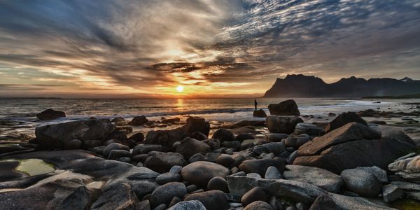 Sunset over a rocky beach with distant mountains and a person standing near the shore.