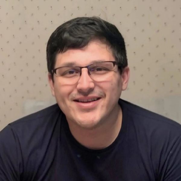 Smiling man wearing glasses and a dark shirt against a patterned wall.