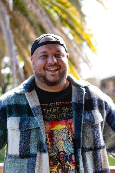 A man with a small beard, green jacket, backwards black cap and a black shirt with Indigenous design
