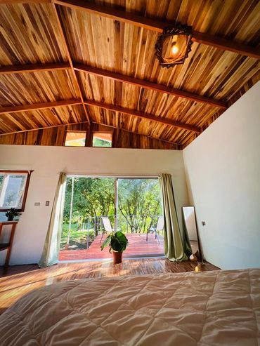 Bright bedroom with wooden ceiling and large glass doors opening to a green outdoor deck.
