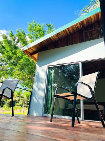 Two chairs on a wooden patio with a modern house and lush greenery under a clear blue sky.
