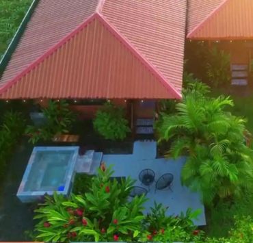 Tropical house with red roof, patio seating, and a small pool surrounded by lush greenery.