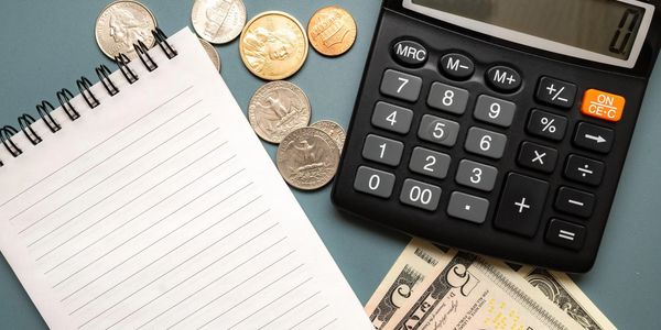 Calculator, coins, bills, and blank notepad on blue surface.