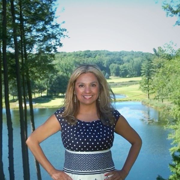 Woman smiling confidently by a lake with trees and hills in the background.