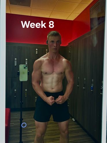 A man in black shorts and no shirt standing in front of lockers.